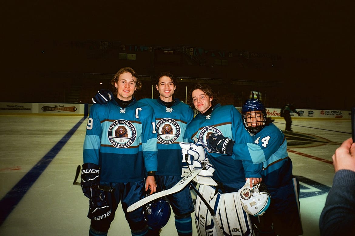 "Four hockey players in blue uniforms posing together on an ice rink, smiling for a photo. The rink is indoors with boards and advertisements visible in the background, suggesting a team game or practice session."