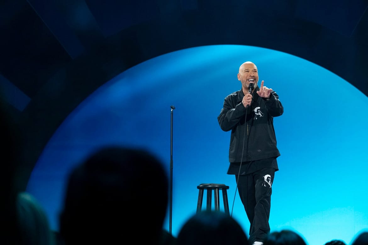 "Comedian performing stand-up on stage, holding a microphone, with a stool nearby and a blue-lit background, engaging a seated audience in a comedy club or theater setting."