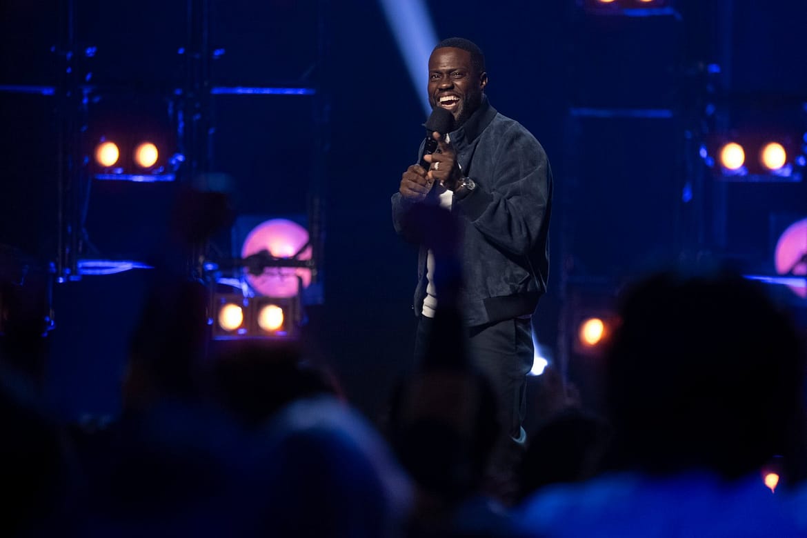 "Man performing stand-up comedy on stage with microphone, smiling and engaging with a cheering audience. Stage is lit with blue and purple lights, background filled with spotlights and stage lighting equipment."