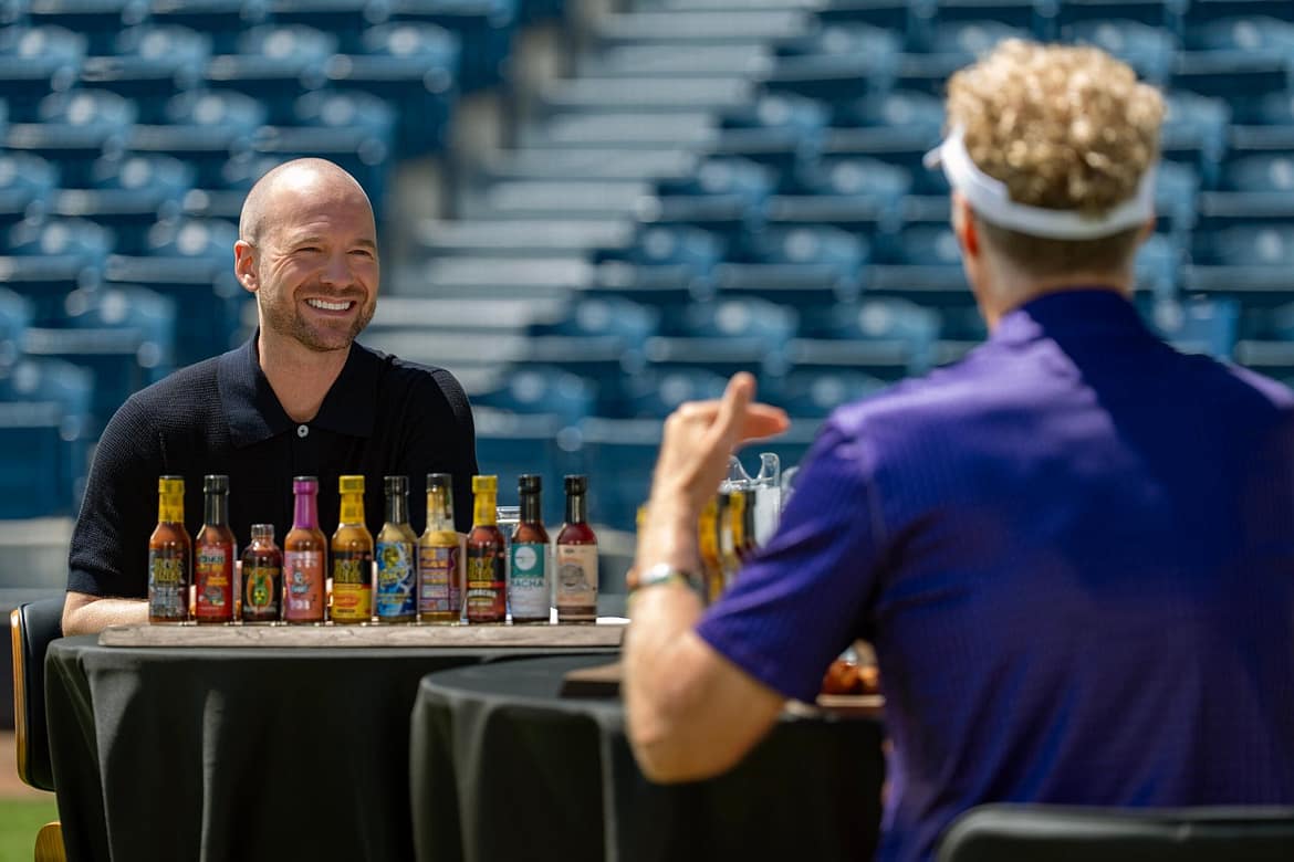 "Two people sit at a table covered with various hot sauce bottles in a stadium with empty blue seats, engaging in conversation during an outdoor event or show."