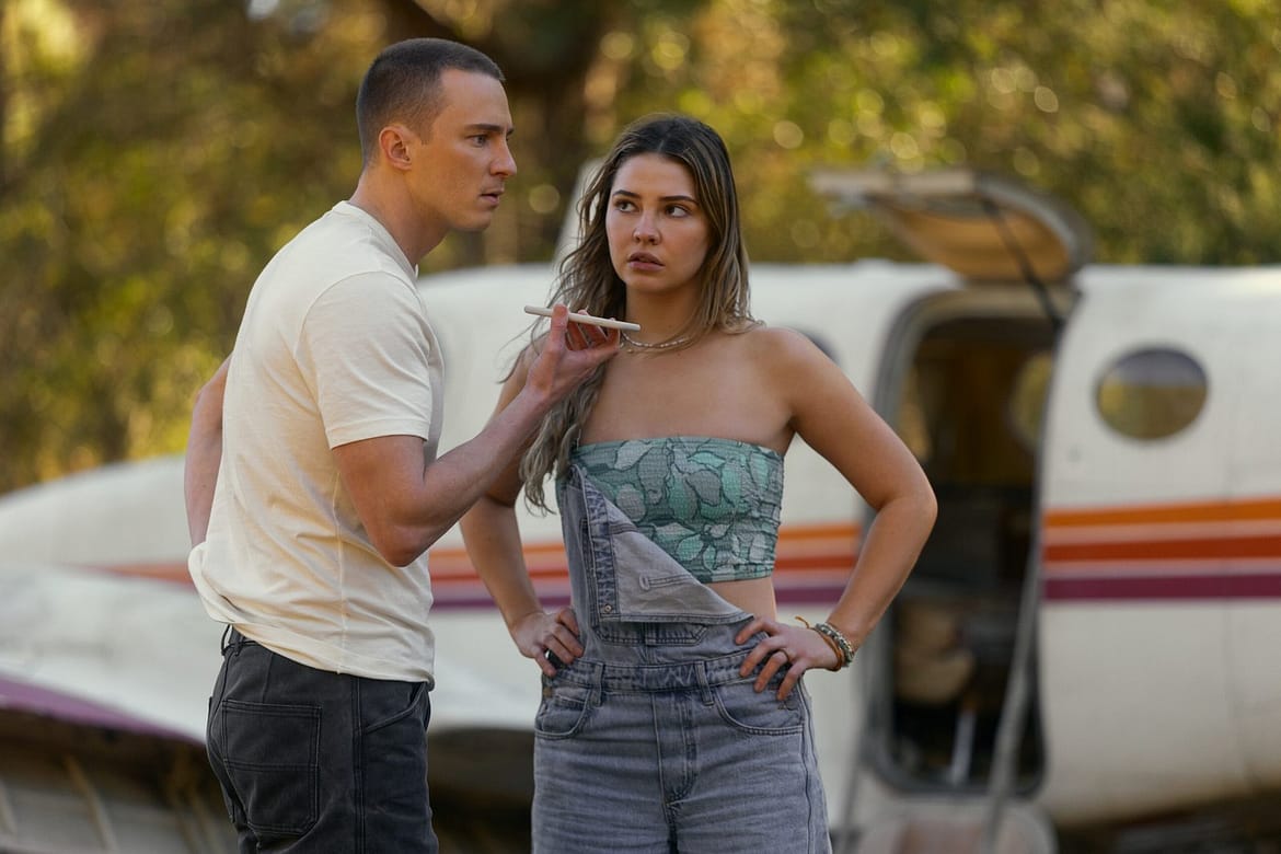 "Two people stand in front of a small plane in an outdoor setting, appearing to be in a serious conversation, with one gesturing while the other listens with arms on hips, surrounded by green trees."