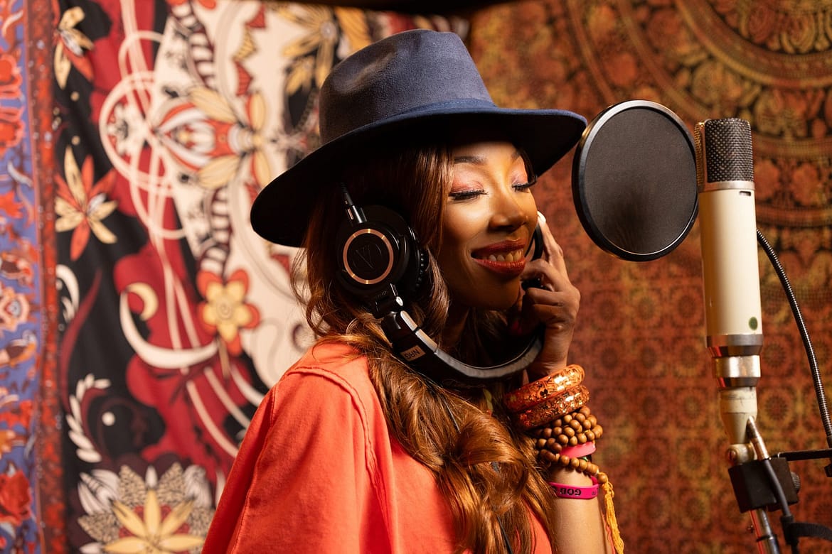 "Woman wearing a hat and headphones singing into a studio microphone, smiling, with colorful patterned tapestries in the background, suggesting a creative, artistic music recording environment."